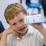 A young student sits in a classroom holding a mobile phone and smiling for a photo.