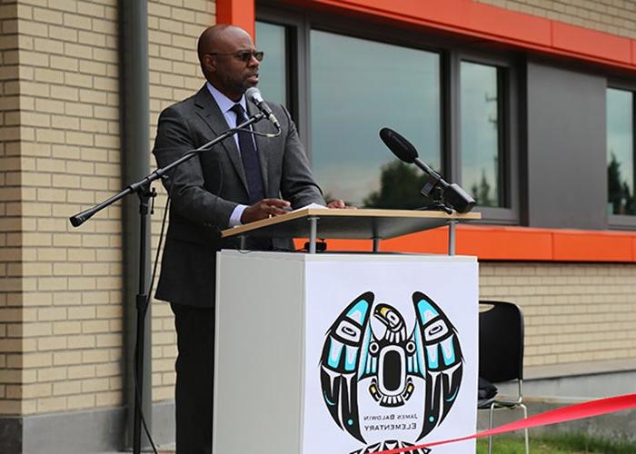 a man stands at a podium with a light colored brick building in the background